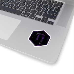 Op de afbeelding: Een paarse en zwarte sticker met een witte rand, in een onregelmatige zeshoekige vorm. De sticker is geplaatst op een zilveren laptop. Het ontwerp van de sticker lijkt een abstracte afbeelding te zijn.