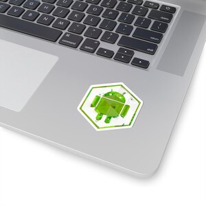 May include: A green Android robot sticker with a white border and a hexagonal shape. The sticker is placed on a laptop. The robot has a 3D appearance with a glossy finish.