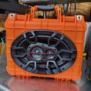 May include: An orange, rectangular speaker with a black oval speaker grill. The speaker has a handle on top and is designed to be portable. The speaker grill has a red accent around the speakers and the brand name "DS18" is visible.