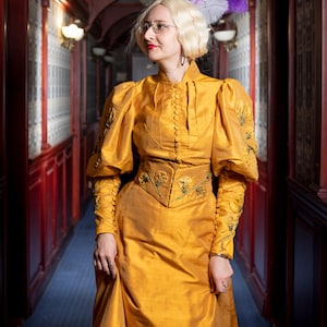 Puede incluir: Un vestido de color amarillo mostaza de manga larga con cuello alto, una hilera de botones en la parte delantera y un panel ajustado en la cintura. El vestido presenta hombros abullonados y detalles florales bordados en las mangas y la cintura. Accesorio de plumas moradas.