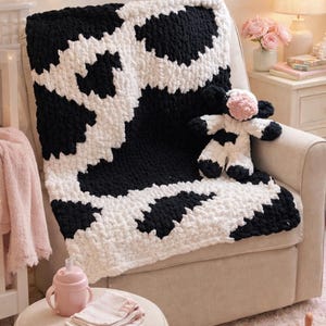May include: A black and white cow print blanket draped over a beige armchair. A matching stuffed cow toy with a pink nose sits on the blanket. A pink sippy cup, burp cloths, and baby shoes are on a small ottoman.