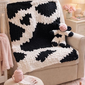 May include: A black and white cow print blanket draped over a beige armchair. A matching stuffed cow toy sits on the blanket. A pink sippy cup, burp cloths, and baby shoes are on a small ottoman.