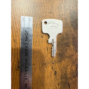 May include: A silver-colored key with a rectangular head and a toothed blade. The key's head has a small hole and an embossed logo. A ruler with the text "JOHN CRANE-HOUDAILLE, INC" is next to the key.