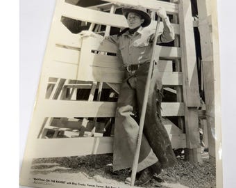 Op de afbeelding: Een zwart-wit vintage foto van een persoon in cowboykleding, inclusief hoed, shirt met lange mouwen en chaps, leunend tegen een houten hek. De tekst onderaan luidt RHYTHM ON THE RANGE met de namen van de castleden.
