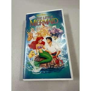 Op de afbeelding: Een VHS-band van Disney's animatiefilm "De Kleine Zeemeermin". De hoes toont Ariel, Prins Eric en andere personages tegen een turquoise achtergrond. De titel is in goud en blauw, met Japanse tekst eronder.