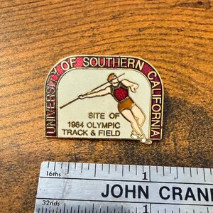 May include: A vintage pin featuring the text "University of Southern California" and "Site of 1984 Olympic Track & Field." The pin depicts a track and field athlete throwing a javelin. The pin is gold-toned with red and white accents.