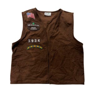 May include: A brown sleeveless vest featuring a Girl Scouts USA Greater Los Angeles patch, a United States flag patch, a numerical patch reading 3934, and a colorful rainbow arch patch. The vest includes a white brand label at the inner neckline.