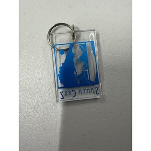 May include: A rectangular clear acrylic keychain with a silver metal ring. The keychain features a blue graphic of a silhouette of a mountain range, a satellite dish, and the text "SANTA CRUZ" in reverse. The number "2" is also visible.