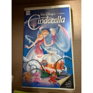 May include: A VHS tape of Walt Disney's animated classic, Cinderella. The cover features Cinderella in a pink dress, the Fairy Godmother in a blue gown, and a pumpkin carriage. The title is in white script on a blue background.