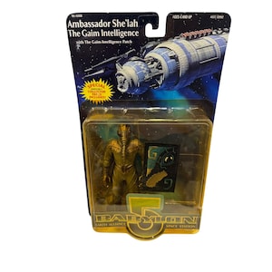 May include: A Babylon 5 action figure of Ambassador She'lah The Gaim Intelligence in original packaging. The card features a space station graphic and text, while the blister pack contains the gold-colored figure and a Gaim Intelligence patch.