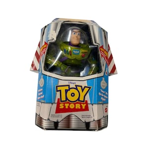 Puede incluir: Una figura de acción de Buzz Lightyear en su empaque original. La caja presenta el logotipo de Toy Story, detalles a rayas rojas y blancas, y texto que indica una altura aproximada de 23 cm. La figura viste un traje verde, blanco y morado.