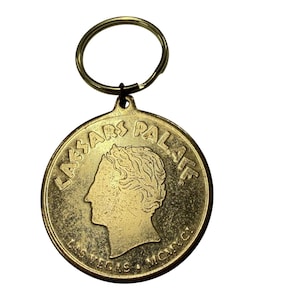May include: A round gold-toned metal keychain featuring a profile relief of a person facing left. The text CAESARS PALACE is embossed at the top, with LAS VEGAS and MCMXC at the bottom. A circular metal split ring is attached to the top of the medallion.
