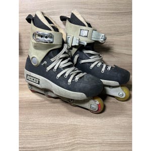 May include: Pair of Roces inline skates. The skates are primarily black with gray and beige accents. They have white laces, gray wheels, and a buckle closure system. The brand name "Roces" is visible on the side.