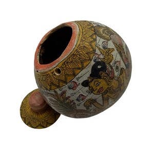 May include: A round, hand-painted container with a matching lid, featuring intricate yellow, black, and white patterns. The exterior displays detailed illustrations of figures and foliage. The container has a wide, open top and a small circular hole.
