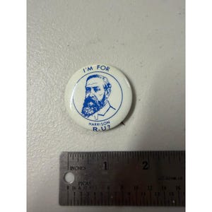 Puede incluir: Un botón redondo blanco con un dibujo lineal azul de un hombre con barba y el texto I'M FOR HARRISON R-U?, colocado sobre una regla metálica que muestra que el botón tiene un diámetro de aproximadamente 3,2 cm.