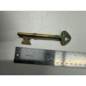 May include: Vintage brass key with a triangular opening in the key head. The key is approximately 5 inches long and lies next to a metal ruler. The key shows signs of age and wear, with a slight green patina.