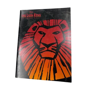 Könnte beinhalten: Ein rechteckiges Programmheft für das Broadway-Musical The Lion King. Das Cover zeigt einen schwarzen Hintergrund mit einer großen, stilisierten Löwengesicht-Grafik in Rot- und Orangetönen. Der Text lautet Disney Presents THE LION KING THE BROADWAY MUSICAL.