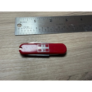 以下が含まれることがあります： 「BUCYRUS-ERIE」と書かれた白いロゴが付いた、小さな赤いポケットナイフ。ナイフの長さは約7.6cmです。インチとパイカの測定値が記載された金属製の定規が背景にあります。