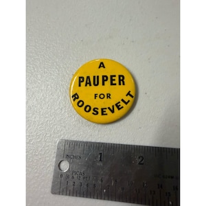 May include: A round yellow pinback button with black text that reads A PAUPER FOR ROOSEVELT. The button is positioned above a metal ruler that shows the item has a diameter of approximately 1.5 inches. The button is circular and flat.