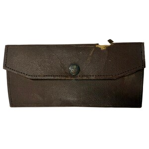 May include: A rectangular dark brown leather wallet featuring a fold-over flap with a central circular metal snap button. The button is embossed with the text Bausch & Lomb. The wallet shows visible wear and surface damage along the top edge.