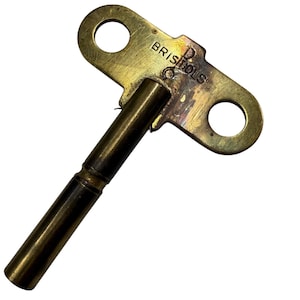 May include: A brass-colored metal clock winding key featuring a T-shaped handle with two circular cutouts. The handle is stamped with the text BRISTOLS. The key has a long, cylindrical shaft with a visible horizontal groove near the center.