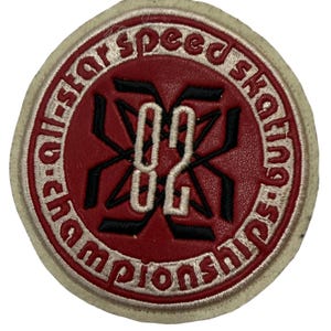 Pode incluir: Um emblema bordado circular com um centro de aspeto de couro vermelho apresentando o número 82 em bordado branco sobre um gráfico de bastões cruzados pretos. A borda é de cor creme com o texto all-star speed skating championships.