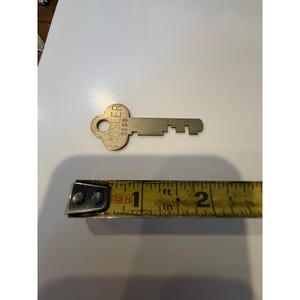 May include: A vintage Mosler 3608 key made of metal. The key is lying on a white surface next to a yellow measuring tape. The key is approximately 2 inches long.