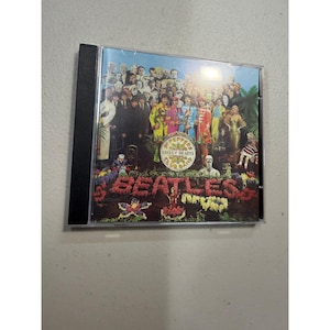 Puede incluir: Una caja de CD con la portada del álbum "Sgt. Pepper's Lonely Hearts Club Band" de The Beatles. La portada muestra una multitud de figuras con atuendos coloridos, con el nombre de la banda y el título del álbum en un arreglo floral.