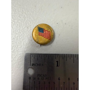 Puede incluir: Un pin circular de tono dorado con una bandera estadounidense impresa. El pin está colocado sobre una regla de metal que muestra que el objeto tiene un diámetro de aproximadamente 1,9 cm. La superficie está desgastada y presenta textura.