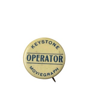 May include: A round, off-white pinback button featuring dark blue text. The word KEYSTONE is arched at the top, the word OPERATOR is centered between two horizontal lines, and the word MOVIEGRAPH is arched along the bottom edge of the button.
