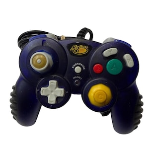 May include: A dark purple Mad Catz video game controller featuring a black cord, a gold-colored left analog stick, a yellow right C-stick, a green A button, a red B button, white X and Y buttons, a white D-pad, and a Start/Pause button.