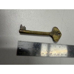 Może przedstawiać: Zabytkowy mosiężny klucz z główką w kształcie rombu i prostokątnym trzonkiem. Klucz ma około 10 cm długości. Metalowa linijka z oznaczeniami cali leży obok klucza.