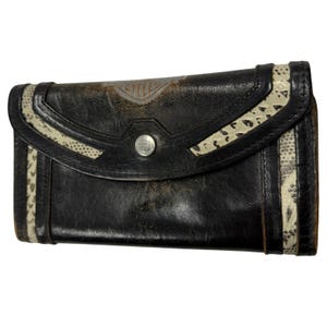May include: A black leather wallet featuring a curved flap with a silver-tone snap button closure. The wallet is accented with cream-coloured textured panels on the sides and flap, and displays a faded Harley-Davidson Cycles logo on the front.