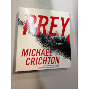 May include: Book cover for Michael Crichton's novel, "PREY". The title is in bold red letters, with the author's name below in red. The cover features a white background with a black splatter design.