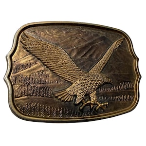 May include: A rectangular brass belt buckle with rounded corners featuring a raised relief design of an eagle in flight. The background depicts a mountain range and a forest of pine trees, all rendered in a metallic gold and dark bronze finish.