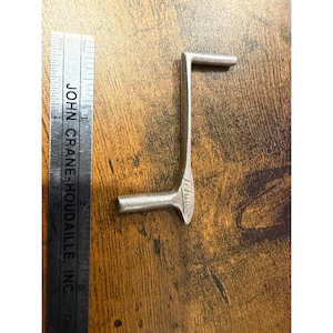 Puede incluir: Una herramienta metálica plateada en forma de L con la palabra "Schuces" grabada en el lateral. Una regla con el texto "JOHN CRANE-HOUDAILLE, INC." y medidas en pulgadas está al lado de la herramienta. La herramienta parece ser un artículo vintage o antiguo.