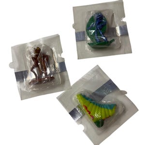 Puede incluir: Tres pequeñas figuras de insectos de plástico selladas individualmente en envases de plástico transparente. Una es un saltamontes marrón, otra es una mantis azul sobre una hoja verde y otra es una oruga segmentada amarilla, verde y roja.