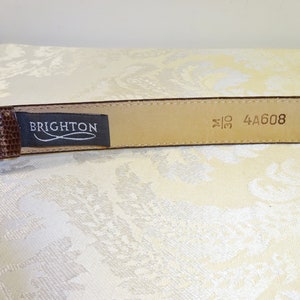 Może przedstawiać: Brązowy sk&oacute;rzany pasek z teksturowanym wzorem przypominającym sk&oacute;rę gada. Pasek ma srebrną klamrę i etykietę z napisem "BRIGHTON". Rozmiar paska to 76 cm.