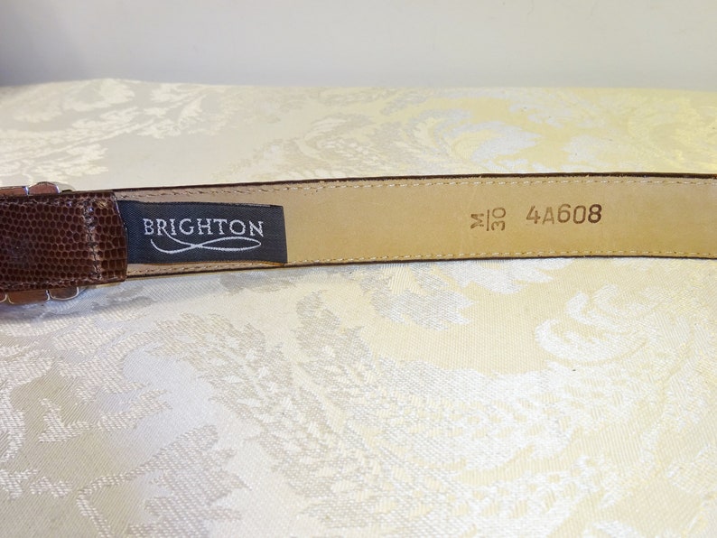 Może przedstawiać: Brązowy sk&oacute;rzany pasek z teksturowanym wzorem przypominającym sk&oacute;rę gada. Pasek ma srebrną klamrę i etykietę z napisem "BRIGHTON". Rozmiar paska to 76 cm.