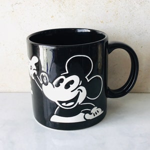May include: Black ceramic mug with a white silhouette of Mickey Mouse. Mickey is smiling and waving with his right hand.