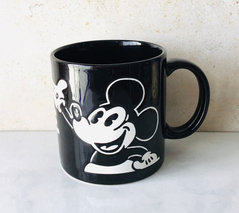 May include: Black ceramic mug with a white silhouette of Mickey Mouse. Mickey is smiling and waving with his right hand.