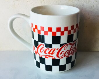 May include: White ceramic mug with a black and red checkered pattern and the Coca-Cola logo in red.