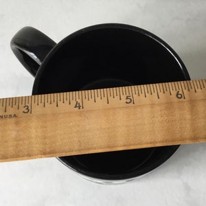 May include: Black ceramic mug with a white handle. The mug is measured with a ruler, showing it is approximately 5 inches wide.
