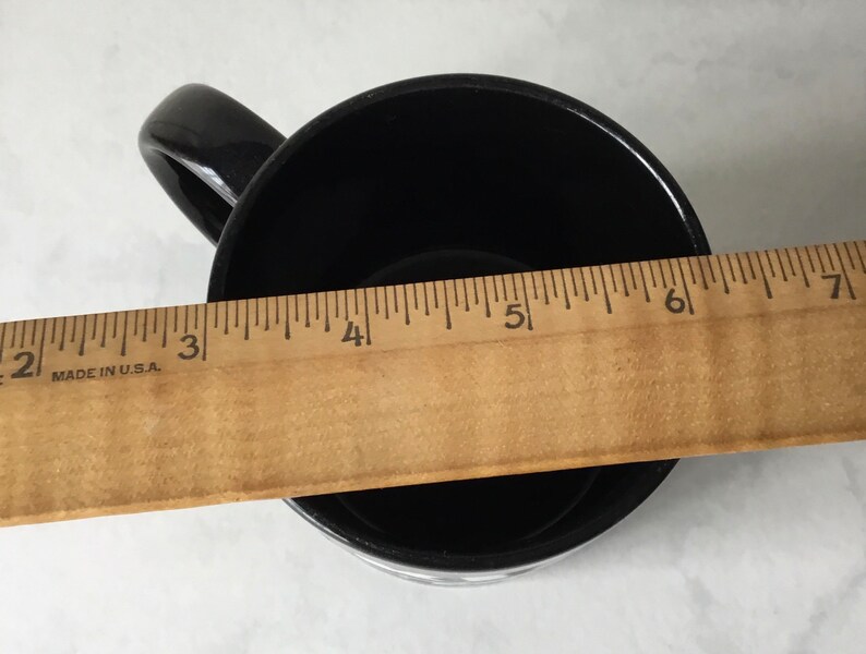 May include: Black ceramic mug with a white handle. The mug is measured with a ruler, showing it is approximately 5 inches wide.