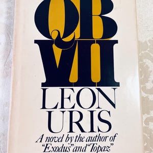 May include: A vintage book cover for "QB VII" by Leon Uris. The title is in large, bold, navy blue and gold lettering. The author's name is in navy blue. The cover is cream-colored with the text "A novel by the author of Exodus and Topaz."
