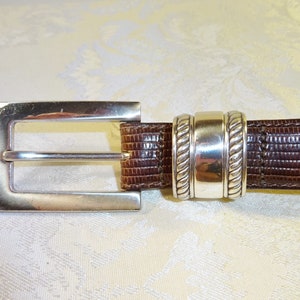 Może przedstawiać: Brązowy sk&oacute;rzany pasek z prostokątną, srebrną klamrą. Pasek ma teksturowany wz&oacute;r i srebrną pętlę w pobliżu klamry. Klasyczny dodatek.