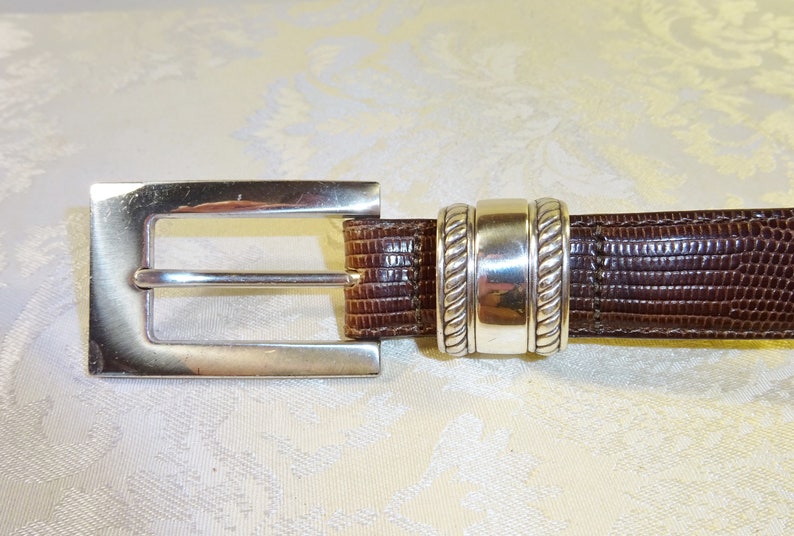 Może przedstawiać: Brązowy sk&oacute;rzany pasek z prostokątną, srebrną klamrą. Pasek ma teksturowany wz&oacute;r i srebrną pętlę w pobliżu klamry. Klasyczny dodatek.