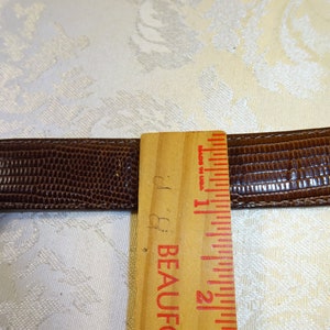 Może przedstawiać: Brązowy sk&oacute;rzany pasek z teksturowanym wzorem przypominającym sk&oacute;rę gada. Drewniana linijka pokazuje pomiary w calach. Pasek ma około 2,5 cm szerokości.