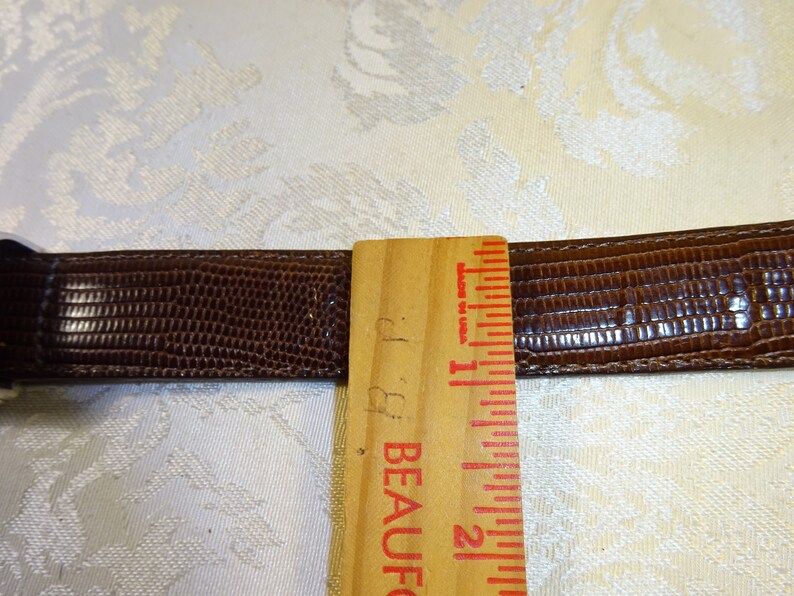 Może przedstawiać: Brązowy sk&oacute;rzany pasek z teksturowanym wzorem przypominającym sk&oacute;rę gada. Drewniana linijka pokazuje pomiary w calach. Pasek ma około 2,5 cm szerokości.