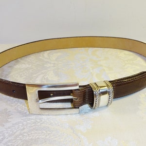 Może przedstawiać: Brązowy sk&oacute;rzany pasek ze srebrną klamrą. Pasek ma teksturowaną powierzchnię i jasnobrązowe wnętrze. Klamra jest prostokątna ze srebrnym wykończeniem. Klasyczny dodatek.